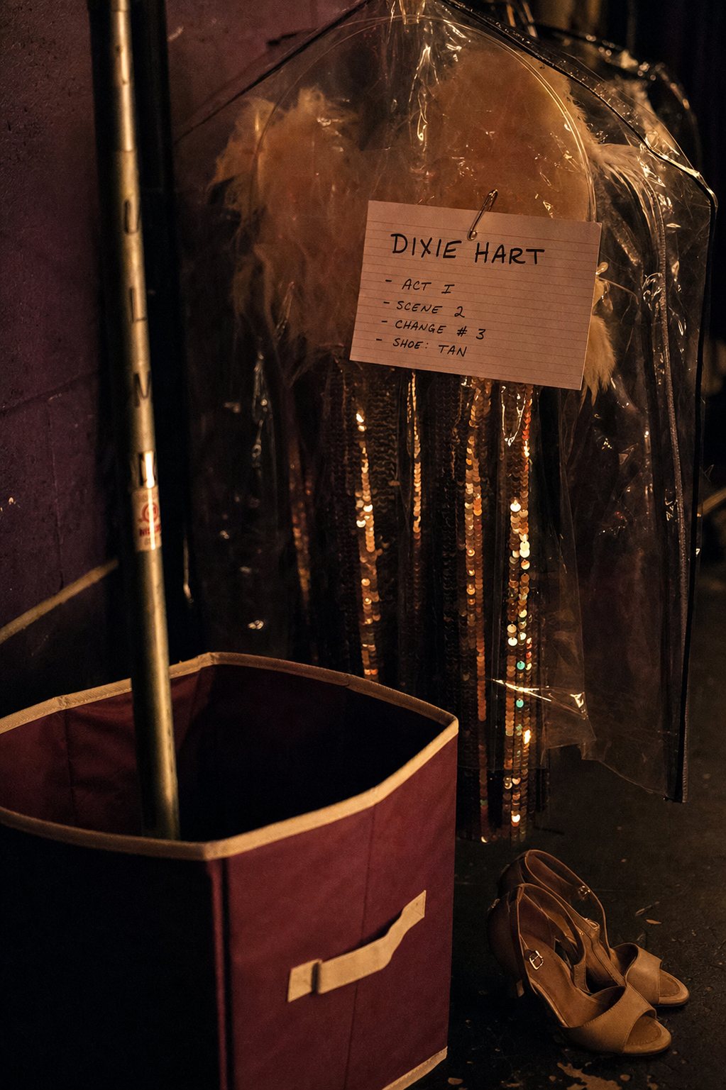 A labeled garment bag on a backstage rack beside a fabric bin and dance shoes.